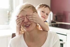 un garçon mignon couvre les yeux de sa mère dans la cuisine photo