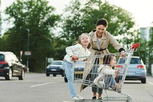 heureuse mère et ses filles s'amusent avec un panier sur un parking à côté d'un supermarché. photo