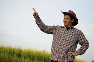 beau fermier asiatique est sur les terres agricoles, porte un chapeau et une chemise à carreaux, met la main sur la hanche et pointe le doigt vers le haut, se sent confiant. copiez l'espace d'ajout de texte ou de publicité. photo