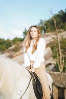 Jeune femme aux cheveux longs en chemise blanche à cheval blanc sur fond de paysage marin photo