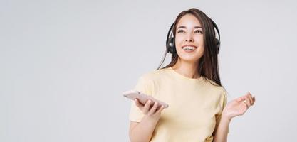 jeune femme asiatique heureuse aux cheveux longs en chemise jaune et jeans utilisant un téléphone portable écoutant de la musique au casque sur fond gris, bannière photo