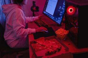 femme gamer ou développeur de logiciels assis devant l'ordinateur personnel moderne et mangeant de la malbouffe la nuit dans la chambre avec des néons photo