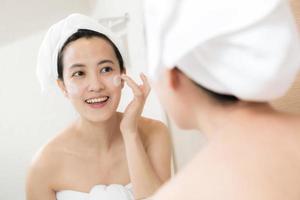 heureuse jeune femme asiatique appliquant des lotions pour le visage tout en portant une serviette et en touchant son visage dans la salle de bain photo