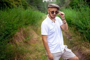 portrait d'un magnifique jeune homme modèle en chapeau et lunettes de soleil regardant loin le parc verdoyant photo