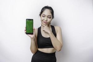 une jeune femme sportive réfléchie porte des vêtements de sport tout en tenant son menton et en montrant un écran vert sur son téléphone, isolée sur fond blanc photo