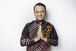 jeune homme asiatique souriant portant une chemise batik, faisant des gestes de salutation traditionnelle isolés sur fond blanc photo