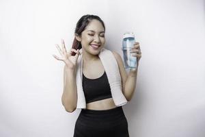 femme asiatique sportive souriante posant avec une serviette sur son épaule et tenant une bouteille d'eau tout en faisant signe ok avec son doigt, se relaxant après l'entraînement photo