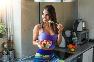 portrait d'une jolie femme souriante caucasienne mangeant de la salade. jeune et heureuse femme mangeant une salade saine assise sur la table avec des ingrédients frais verts à l'intérieur photo