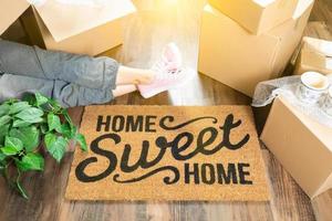 femme portant des sueurs relaxantes près du tapis de bienvenue home sweet home, des boîtes de déménagement et des plantes. photo