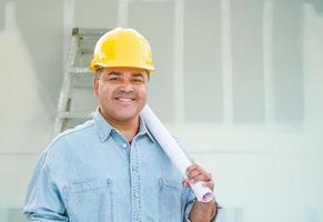 entrepreneur masculin hispanique avec des plans de plan directeur portant un casque de protection devant les cloisons sèches et l'échelle photo