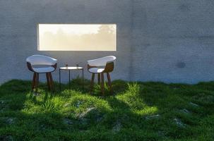 deux chaises, une table et des verres à eau près de la fenêtre avec la lumière du soleil qui brille à travers le mur de béton dans le champ d'herbe et de rochers. photo