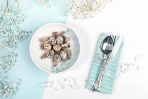 table de fête de la pâque avec assiette avec œufs de caille, fleurs blanches, couverts sur serviette sur fond blanc et bleu clair. photo