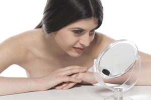belle femme brune séduisante vérifiant la peau de son visage devant un miroir sur fond blanc. photo