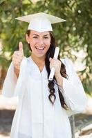 mixed race thumbs up girl célébrant l'obtention du diplôme à l'extérieur en bonnet et robe avec diplôme en main photo