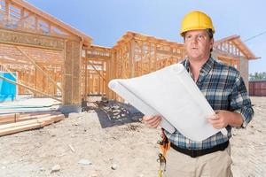 entrepreneur masculin avec des plans de maison portant un casque en face de l'ossature de la construction d'une nouvelle maison photo