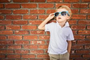 mignon jeune garçon caucasien portant des lunettes de soleil contre le mur de briques photo