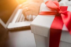 coffret cadeau blanc avec ruban rouge et arc près de l'homme tapant sur un ordinateur portable. photo