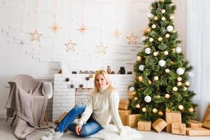 photo de mode de noël de belle fille aux cheveux blonds dans des vêtements confortables posant près de l'arbre du nouvel an avec des cadeaux