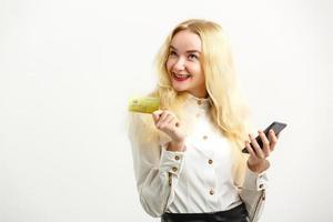 portrait d'une jeune fille blonde heureuse montrant une carte de crédit en plastique tout en tenant un téléphone portable isolé sur fond photo