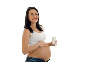 portrait de jeune femme enceinte drôle avec un verre de lait touchant son ventre et souriant à la caméra isolée sur fond blanc photo