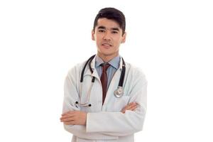 beau jeune homme brune médecin en uniforme blanc avec stéthoscope et mains croisées isolées en studio photo