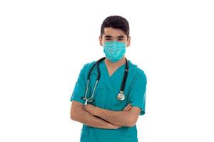 portrait de jeune beau médecin homme brune en uniforme bleu et masque avec stéthoscope regardant la caméra isolée sur fond vide photo