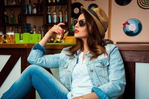 femme élégante en lunettes de soleil et chapeau buvant un cocktail d'alcool froid au bar photo