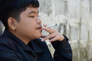 garçon apprenant à fumer par lui-même dans la zone située derrière la clôture de l'école qui est un endroit caché pour les gens, mauvaise influence de la vie au lycée ou au collège, dépendance. photo