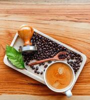 vue de dessus de la tasse à café expresso et de l'équipement de l'outil de café barista inviolable et des grains de café torréfiés foncés avec des feuilles de café vert sur une table en bois photo