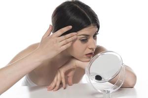 belle femme brune séduisante vérifiant la peau de son visage devant un miroir sur fond blanc photo