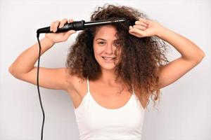 belle femme souriante aux longs cheveux ondulés le repassant, à l'aide d'un fer à friser. fille heureuse avec de magnifiques cheveux lisses et sains utilisant un bigoudi pour des boucles parfaites. coiffure et coiffure photo