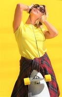 portrait d'une belle fille blonde heureuse tient son skateboard longboard en bois sur un mur de fond jaune, regarde vers la caméra et sourit. scène urbaine, vie urbaine. dame mignonne hipster. photo