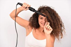 belle femme souriante aux longs cheveux ondulés le repassant, à l'aide d'un fer à friser. fille heureuse avec de magnifiques cheveux lisses et sains utilisant un bigoudi pour des boucles parfaites. coiffure et coiffure photo