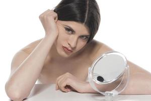 belle femme brune séduisante vérifiant la peau de son visage devant un miroir sur fond blanc. photo