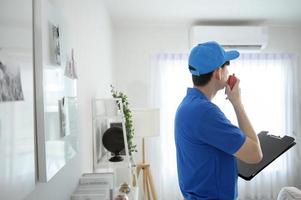 un jeune technicien asiatique homme de service portant un uniforme bleu vérifiant, nettoyant le climatiseur à la maison photo