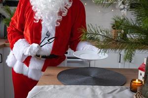 le père noël met la table à la maison dans la cuisine blanche, met une assiette, met une fourchette et un couteau à couverts. préparer la fête de noël, mettre la table pour les invités photo