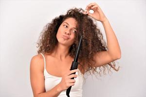 cheveux bouclés. belle femme souriante aux longs cheveux ondulés le repassant, à l'aide d'un fer à friser. fille heureuse avec de magnifiques cheveux lisses et sains utilisant un bigoudi pour des boucles parfaites. coiffure et coiffure photo