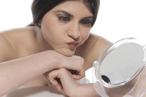 belle femme brune séduisante vérifiant la peau de son visage devant un miroir sur fond blanc. photo