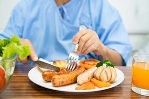 une vieille dame asiatique ou une vieille dame patiente mangeant un petit-déjeuner de steak de saumon avec des aliments sains aux légumes tout en étant assise et affamée sur son lit à l'hôpital. photo