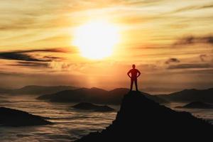 silhouette homme debout et akimbo au sommet d'un rocher de pierre dans la montagne et regardant le brouillard, la brume, le coucher du soleil et la lumière du soleil au lever du soleil. vallée brumeuse. homme randonnée.personne silhouette stand. concept de nature de montagne. photo