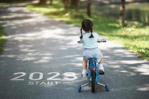 bonne année 2023, 2023 symbolise le début de la nouvelle année. la lettre commence la nouvelle année 2023 sur la petite fille faisant du vélo sur la route dans le jardin du parc naturel. objectif de réussite. fond d'écran numéro 2023. photo