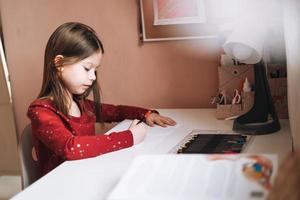 jolie écolière aux cheveux longs en robe rouge dessine avec un crayon à table dans la chambre des enfants à la maison photo