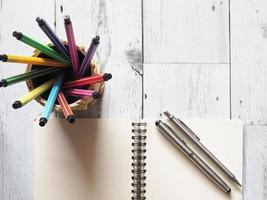 stylo tableau coloré circulaire dans une boîte avec cahier ouvert vierge avec deux stylos argentés sur table en bois blanc et lumière naturelle du concept d'idée de soleil vue de dessus, affaires et éducation photo