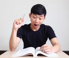 homme asiatique choqué et excité montrer le stylo et regarder le livre sur le bureau sur blanc isolé photo