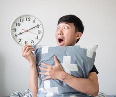 homme asiatique visage choqué et regardant l'horloge à la main et étreignant l'oreiller sur le concept de réveil tardif isolé blanc photo