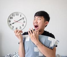 homme asiatique visage choqué et regardant l'horloge dans la main et l'oreiller câlin sur le concept de réveil tardif isolé blanc photo