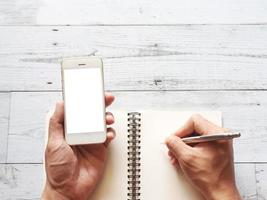 main tenant l'écran blanc du smartphone et écrire sur une page vierge avec un stylo argenté sur une table en bois blanc vue de dessus l'ombre de la nature de l'espace photo
