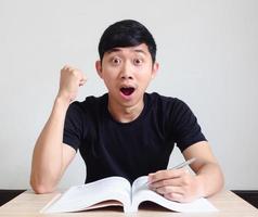 jeune homme asiatique se sent choqué visage excité et poing avec le livre sur le bureau regarde la caméra sur blanc isolé photo