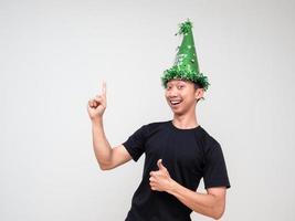 jeune homme asiatique visage heureux regarder la caméra avec un chapeau vert pointer le doigt droit au-dessus du pouce vers le haut et un sourire heureux sur fond blanc isolé photo