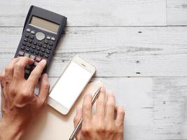 main avec un stylo argenté et un smartphone, appuyez sur le bouton de la calculatrice sur une page vierge de journal sur une table en bois vue de dessus nature shadow copy space business concept photo